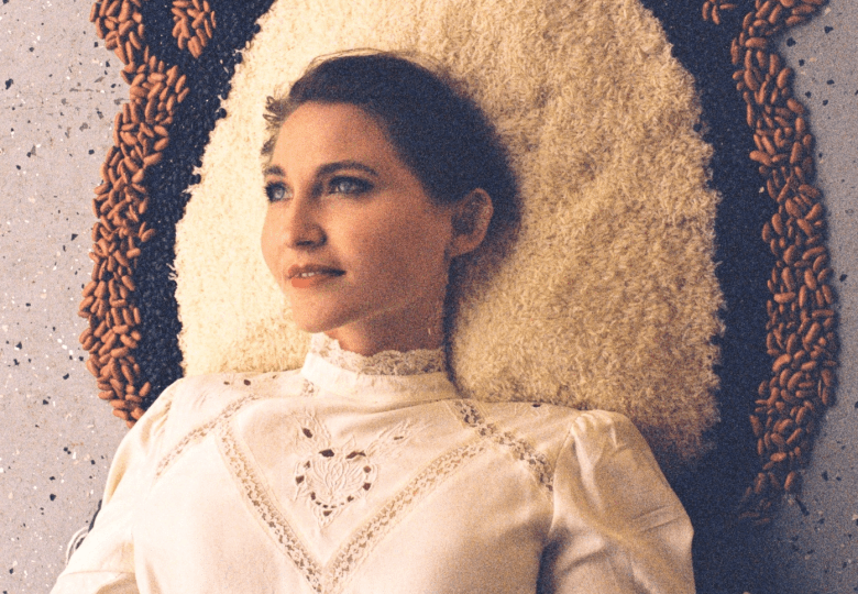 Musician June Bunch lying down against a mosaic background made of rice, black beans, and brown beans arranged in an ornate, halo-like pattern. The person wears a vintage white high-neck lace blouse and looks off to the side with a serene expression.