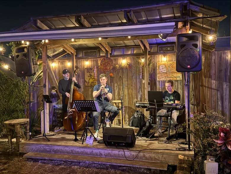 The Dan Calkins Trio performing live on a rustic wooden outdoor stage at New World Tampa. Three musicians are visible: one playing an upright bass, one playing trumpet while seated, and one playing a keyboard. Warm string lights and professional speakers frame the stage under a wooden roof.
