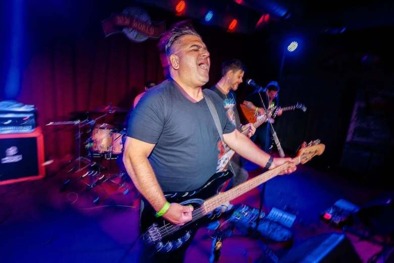 Members of the Tampa band Right On Time performing live on a dark stage with red and blue lighting. In the foreground, a person sings passionately while playing a black bass guitar. A drummer and another guitarist are visible in the background under a 'New World' venue sign.