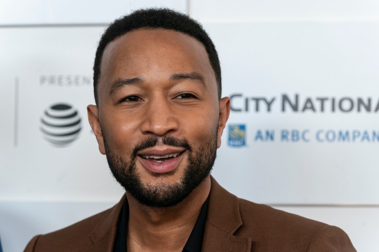 Musician John Legend smiling in a close-up portrait; the person wears a brown blazer over a black shirt. The background features a white press wall with the 'City National Bank' and 'RBC Company' logos.