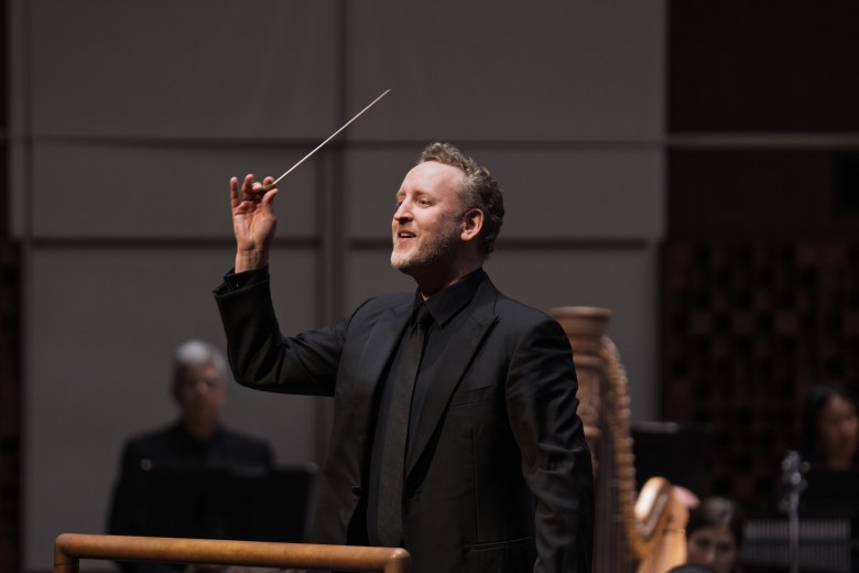 Michael Francis, Music Director of The Florida Orchestra, in a side-profile portrait. The person is smiling and looking upward, wearing a professional black suit, black dress shirt, and a black tie against a neutral grey background.