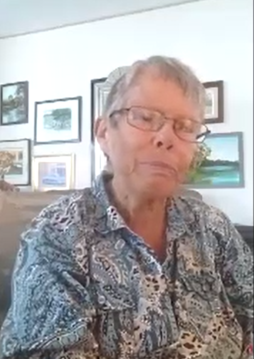 Woman sits in a living room in a blue and white shirt with gray hair and glasses.