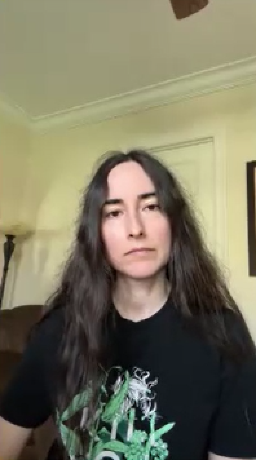 Woman sits in her room in a black shirt with long brown hair.