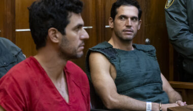 photo of a man in a red jumpsuit and another man wearing a navy kevlar vest inside a court room.