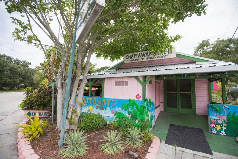 The exterior of The Chattaway, a historic restaurant in St. Petersburg, Florida. The pink wooden building features a mural of pink flamingos and green palm fronds. A white sign reading 'Chattaway Drive-In' sits atop a gabled roof, surrounded by lush tropical plants, succulents, and a large oak tree under a clear sky.