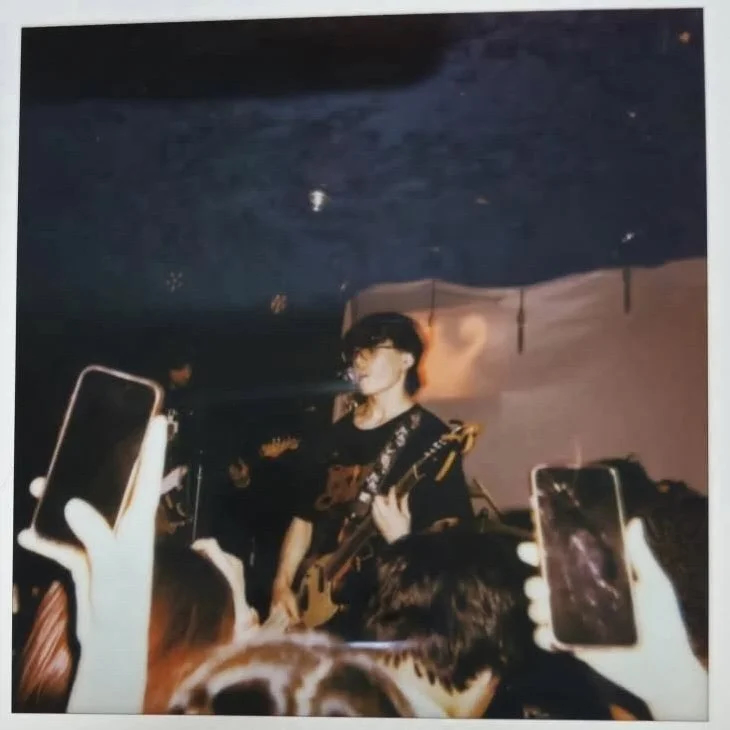 A polaroid-style photo of the band flyingfish performing a live concert. A person with dark hair and glasses plays an electric guitar on a dark stage. In the foreground, the silhouettes of audience members hold up smartphones to record the performance against a backdrop of a simple white sheet and ambient stage lighting.