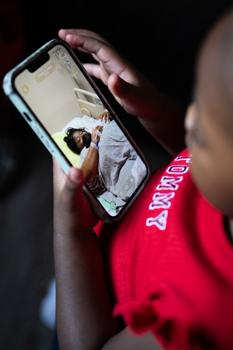 A girl wearing a red “Tommy” shirt looks at a smartphone with a picture of a woman wearing a face mask and lying down in a hospital bed.