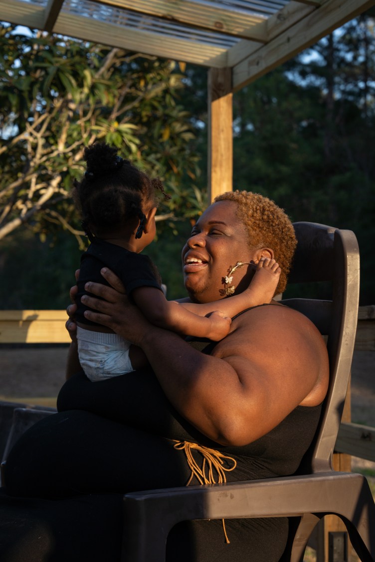 A smiling Black woman sits in a plastic chair under a wooden awning, holding a baby wearing a diaper. Light from a sunset casts a shadow on the baby and lights up the woman.