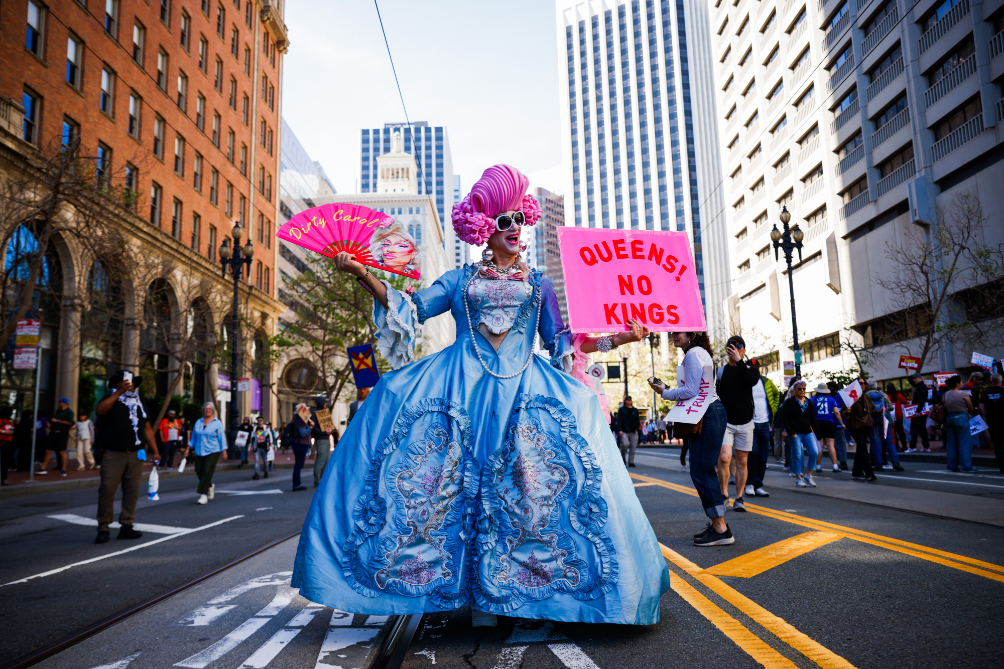 A drag queen is in a big Victorian style dress with fans. One reads: QUEENS! No Kings!