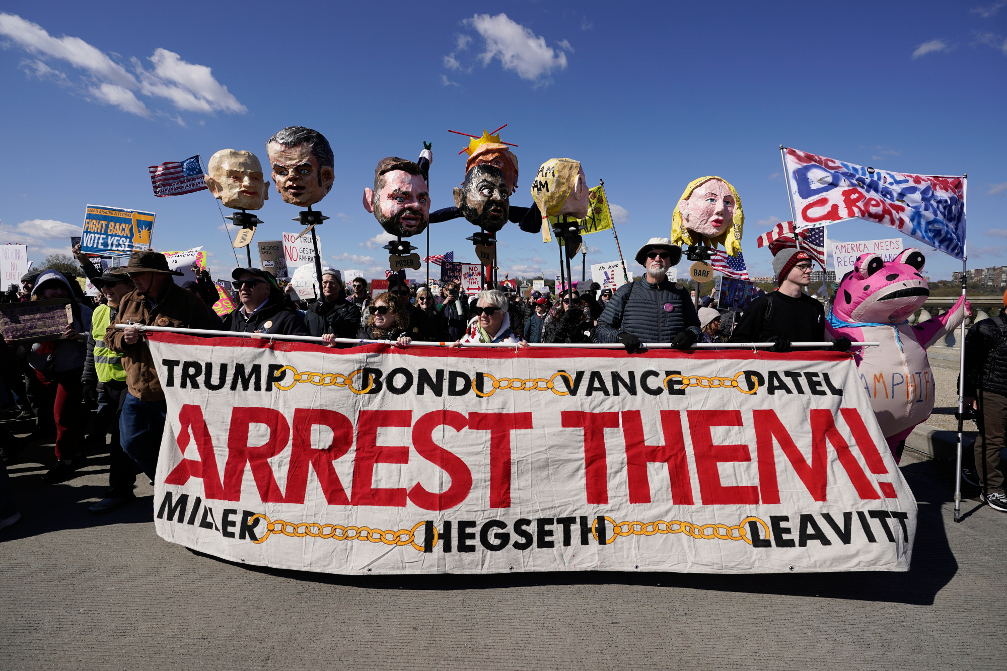 protestors carry depictions of prominent trump admin leaders with a sign that says "arrest them."