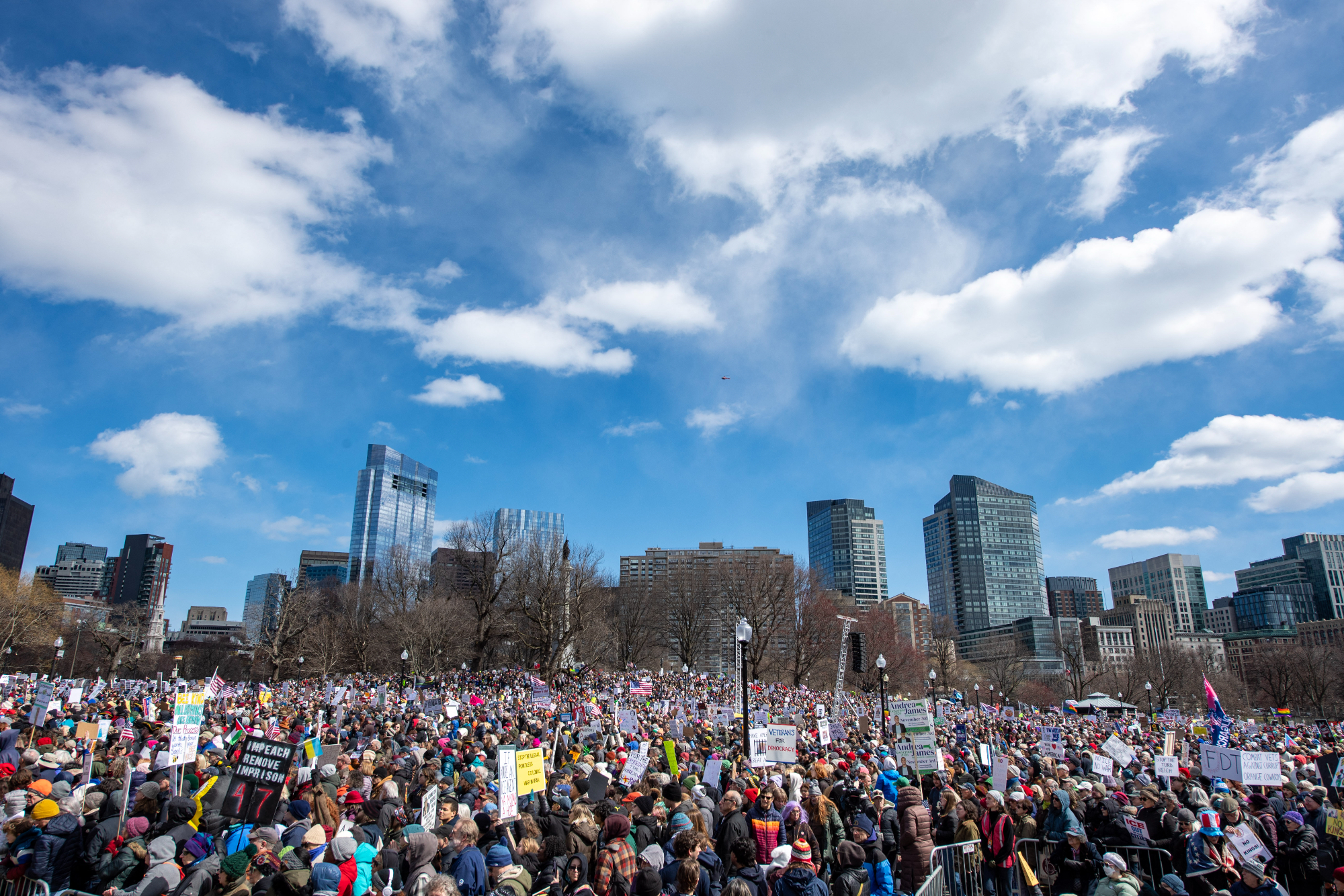 A large crowd gathers and are seen below a blue sky and set against city buildings.