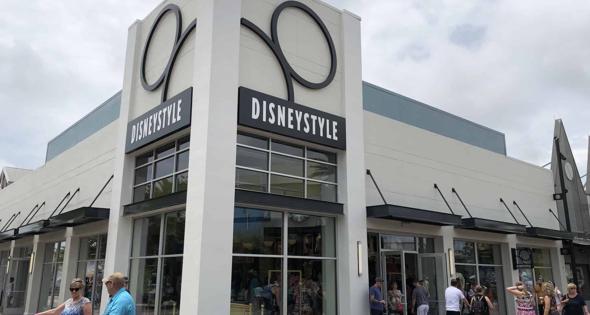 Exterior of the DisneyStyle store at Disney Springs, featuring a large pair of Mickey Mouse ears adorning the facade. Several people walk past and enter the store on a partly cloudy day, possibly eyeing a festive Halloween Belt Bag or charming Shirt Set inside.