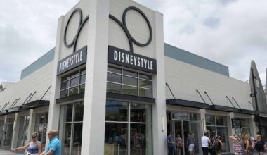 Exterior of the DisneyStyle store at Disney Springs, featuring a large pair of Mickey Mouse ears adorning the facade. Several people walk past and enter the store on a partly cloudy day, possibly eyeing a festive Halloween Belt Bag or charming Shirt Set inside.