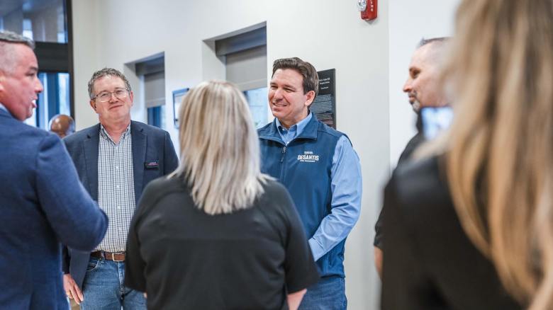DeSantis for President" vest, smiling and talking with a group of supporters during a campaign event.