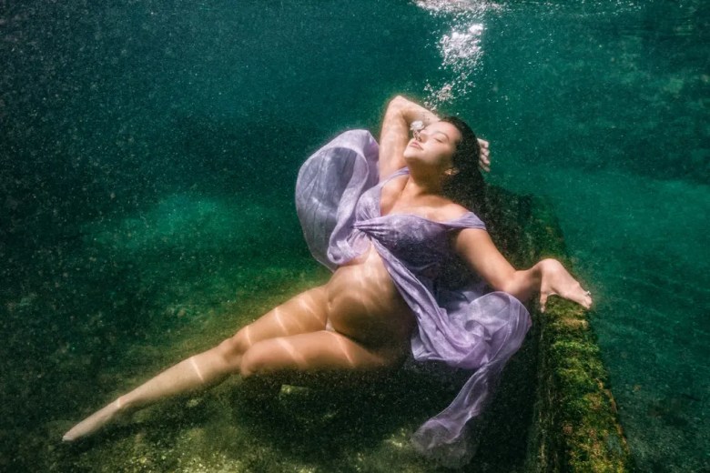 An artistic underwater maternity portrait of a pregnant woman posing gracefully in a flowing lavender dress.