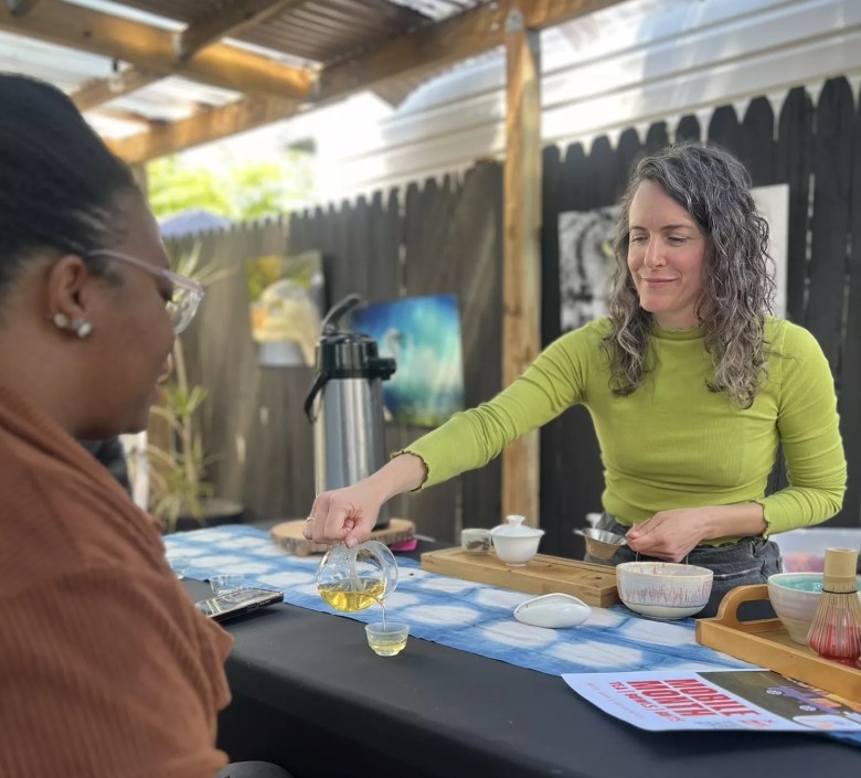 A smiling vendor in a green shirt pours a small tasting cup of tea for a customer at an outdoor market stall.