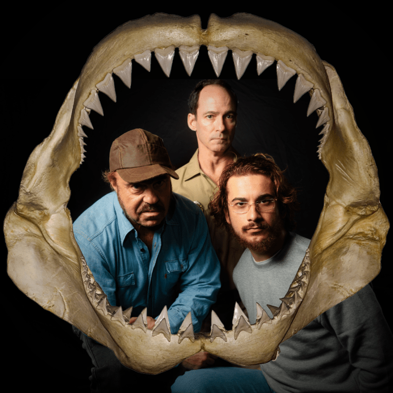 Three actors peer at the camera from inside a shark jaw