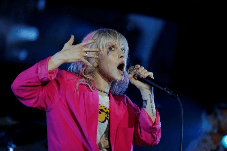 Musician Hayley Williams of Paramore performing live on stage. The person has blonde hair with pink highlights and blue makeup under one eye, wearing a bright pink oversized blazer over a vintage graphic t-shirt. They are singing into a microphone with one hand raised near their face in an expressive gesture.