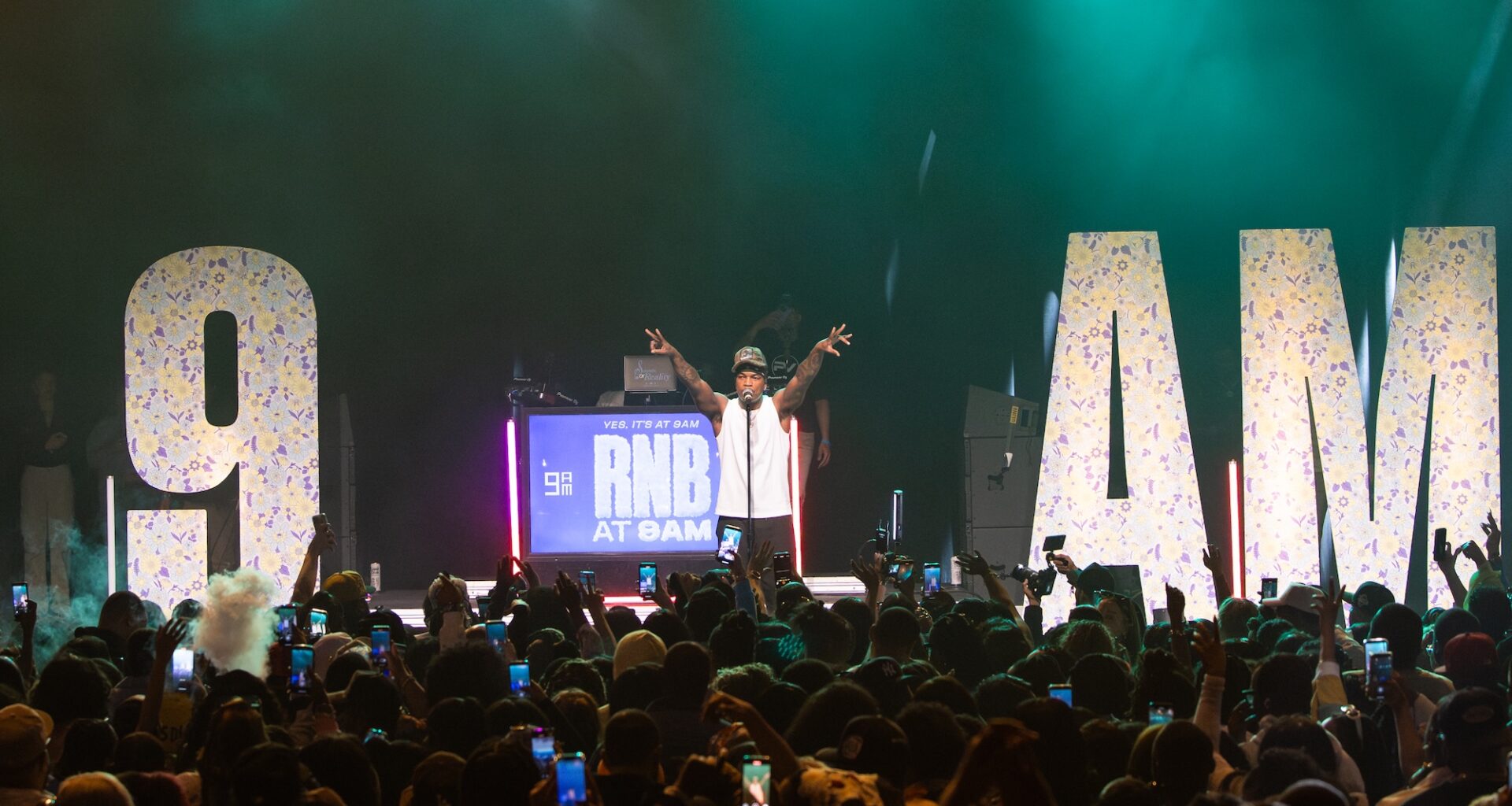 Photo of a man on stage in front of a crowd. The stage has letters that read 9am .
