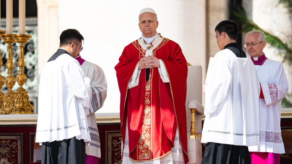 Pope Leo, dressed in a red robe with a white robe underneath, stands with his hands clasped in front of his chest. Leo is standing in front of a white chair. Four other people are standing surrounding him in white clothing.