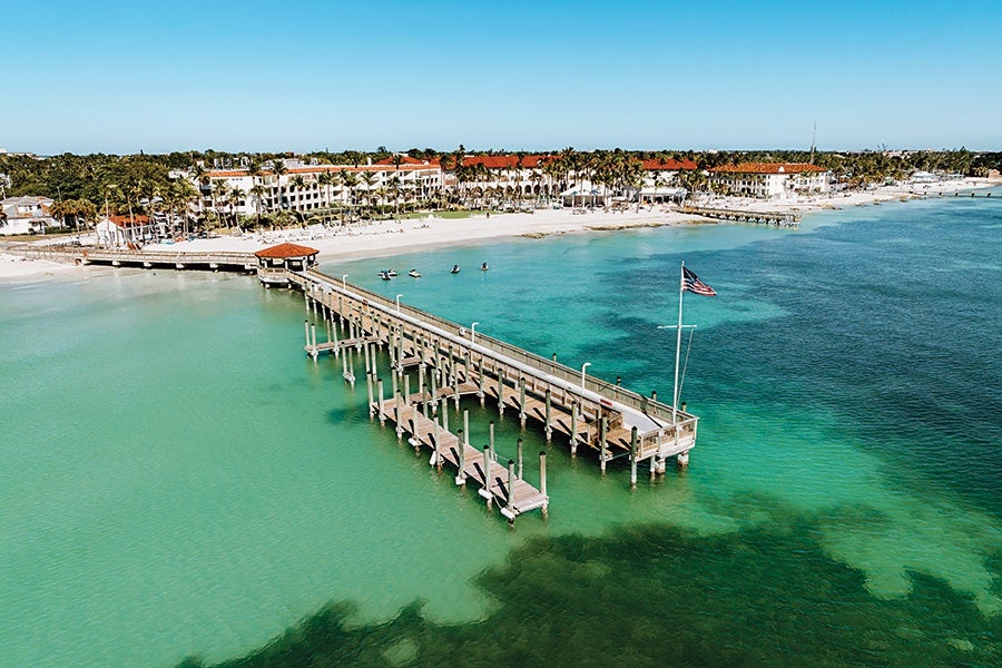 A long wooden pier extends over clear turquoise water toward a sandy beach lined with palm trees and white buildings with red-tiled roofs. An American flag flies at the end of the pier, and a few small boats are near the shore. The sky is clear and blue.