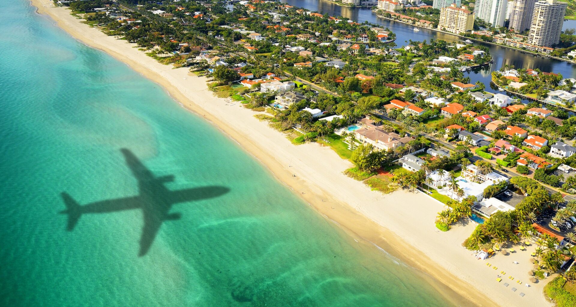 aerial photo of the shadow of a plane flying over the ocean in the direction of trees, houses, and skyscrapers in Miami
