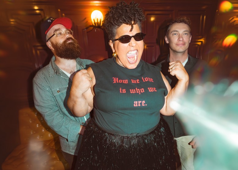 Brittany Howard of Alabama Shakes wearing sunglasses and a black "How we Love is who we are" shirt, posing energetically with bandmates Zac Cockrell and Steve Johnson in a dimly lit room.