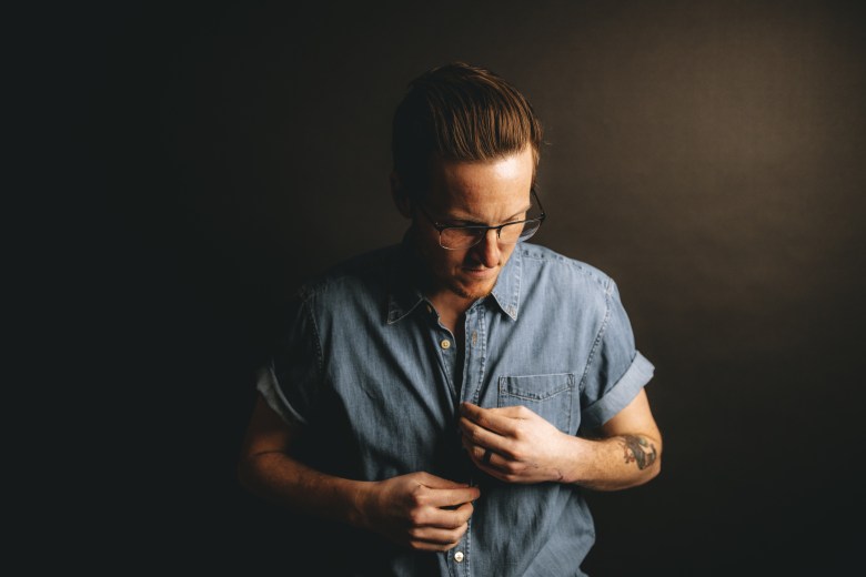Musician Andy Sydow in a moody studio portrait against a dark background; the person wears glasses and a denim shirt with rolled sleeves and looks downward while adjusting a button. A tattoo is visible on the person's left forearm.
