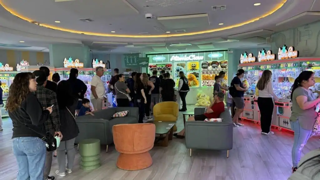 A large group of kids and adults play claw machines inside a circular arcade. Bright yellow and green lights illuminate the machines.