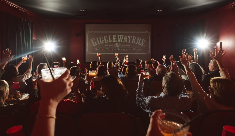 A lively crowd inside Gigglewaters Social Club & Screening Room raises drinks in unison toward the movie screen, which displays the venue’s logo. The dim theater is filled with people cheering, holding cocktails, and celebrating together under the glow of projector lights.