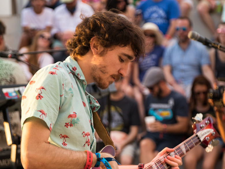 Musician Jaron Jammer performing live, playing a bass guitar while wearing a light blue short-sleeve button-down shirt with a pink flamingo print. The person is in profile, focused on the instrument, with a blurred audience in the background.