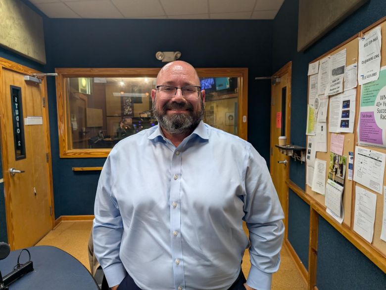 A portrait of Brian Nathan, candidate for Florida State Senate District 14. They are a bald person with a salt-and-pepper beard and glasses, smiling warmly at the camera. They are wearing a light blue button-down shirt and standing in a room with blue walls. In the background, there is a wooden door, a window looking into another room, and a bulletin board with various papers pinned to it.