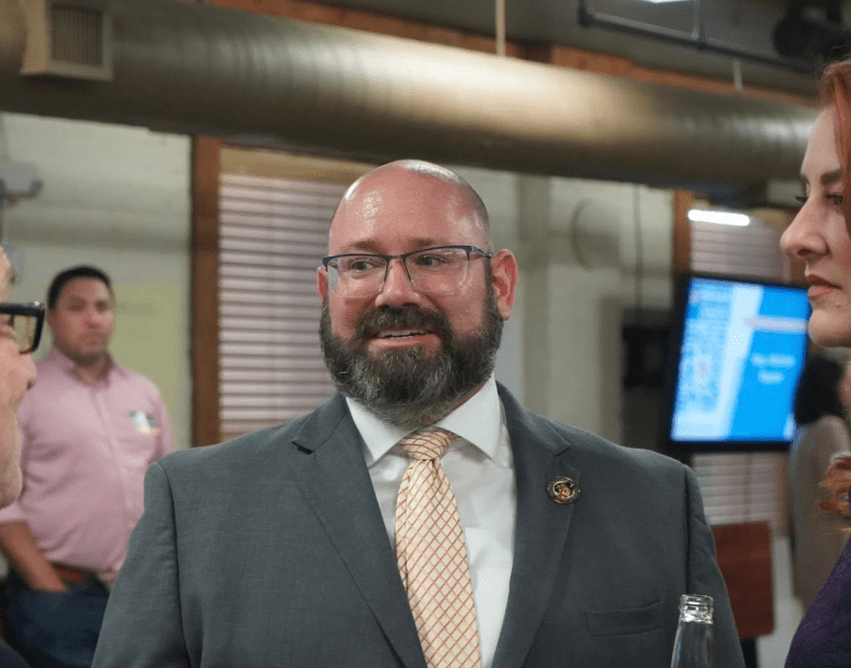 Florida Senate candidate Brian Nathan smiling in a professional indoor setting. The person has a short-cropped beard and glasses, wearing a dark grey suit jacket with a light yellow tie. Other individuals in business attire are visible in the soft-focus background of the office or event space.