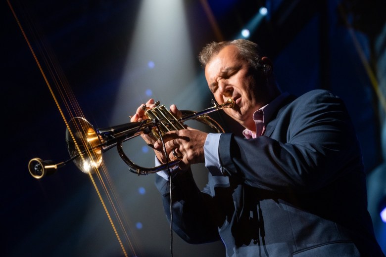 Musician Carl Fischer performing a trumpet solo under a bright spotlight. Fischer is wearing a dark suit jacket and a pink button-down shirt, holding the trumpet with both hands in a focused, emotive pose against a dark stage background.