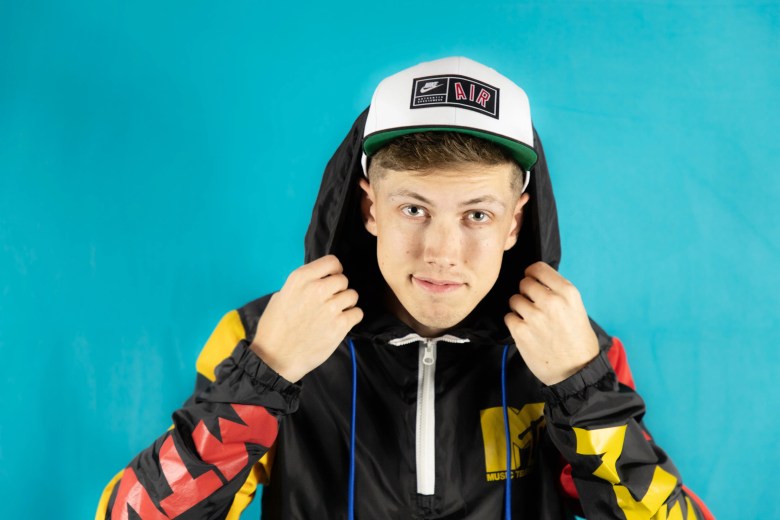 Close-up portrait of comedian Cam Bertrand smiling against a solid turquoise background. The person wears a white baseball cap with a green brim and a black shirt.