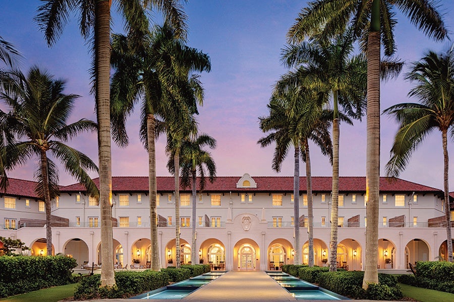 A large, elegant building with white walls and a red-tiled roof, featuring multiple arched doorways and windows illuminated with warm lights. Tall palm trees line a central walkway leading to the entrance, flanked by narrow water features on both sides. The sky is a soft gradient of purple and blue, suggesting dusk.