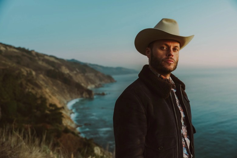 Charley Crockett wearing a cowboy hat and denim jacket, standing on a coastal cliffside at sunset with the ocean in the background.