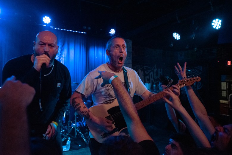 A high-energy live concert photograph of the band Poison The Well performing at Crowbar in Tampa on March 23, 2026. Vocalist Jeffrey Moreira, with a beard and wearing a black t-shirt, shouts into a microphone on the left. Beside them, a guitarist in a white t-shirt plays a light-colored electric guitar with an intense, open-mouthed expression. Several fans' arms are raised in the foreground, reaching toward the stage under blue and white stage lights.