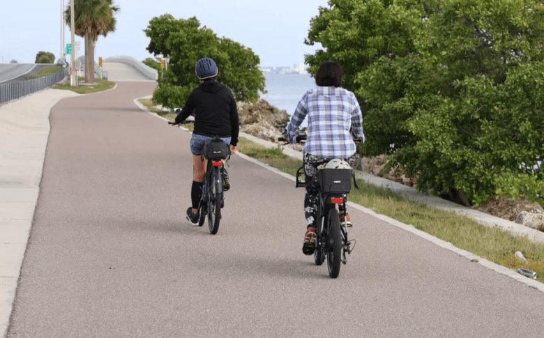 Two cyclists ride along the paved Courtney Campbell Trail in Tampa Bay. The path follows the waterfront with lush green trees and the open bay in the background. Photo courtesy of Visit St. Pete/Clearwater.