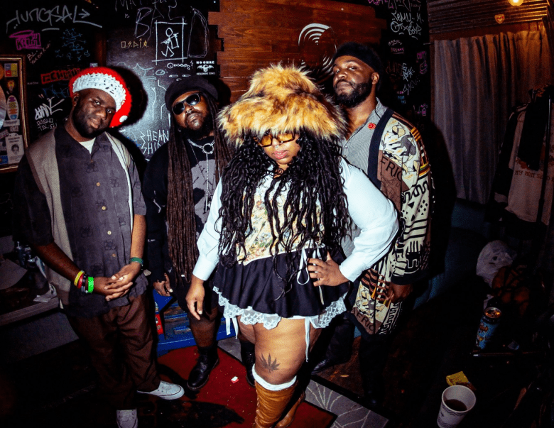 The four members of the band Cozy In The Black pose in a dimly lit room with walls covered in white graffiti and stickers. The central figure wears a large faux-fur trapper hat, a lace-trimmed skirt, and tall brown boots; the other three members stand behind them wearing eclectic headwear, including a red and white knit cap and a black beanie.