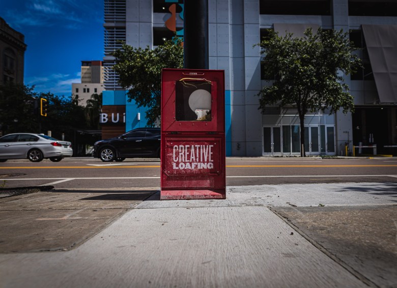 A red Creative Loafing newsstand box standing on a concrete sidewalk in Downtown Tampa. The box is centered in the frame, featuring the 'Creative Loafing' logo in white serif text on its front panel. The background shows a sunlit street intersection with a white car and a black SUV passing by. Modern multistory buildings, including one with 'BUILT' signage, and green trees sit under a clear blue sky. The perspective is low, looking down a wide sidewalk toward the street.
