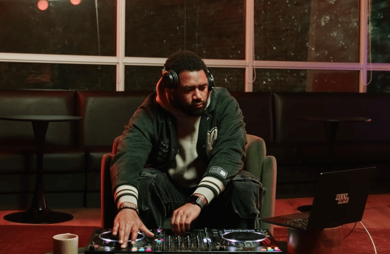 A screen capture of DJ Ryan Rose performing a 'How A Sunset Sounds' set. The person has a dark beard and wears a dark green hoodie with white stripes on the sleeves and large black headphones. They are focused on a silver DJ controller and a laptop on a red surface, with a blurred background of large windows and a dark lounge interior.