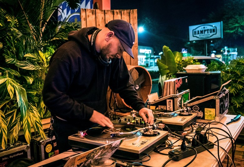 DJ Cub performing a live vinyl set at an outdoor night event. The person wears a black hoodie and a dark baseball cap, leaning over two turntables and a mixer on a table. Lush green plants and a 'Hampton' sign are visible in the background.