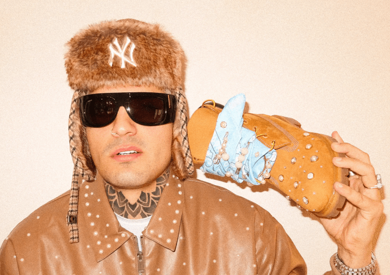 Musician Dale Pututi in a high-fashion portrait; the person wears a brown leather jacket adorned with pearls, a patterned trapper hat, and large dark sunglasses. A blue paisley bandana with pearl accents is tied around a tan boot held near the shoulder.