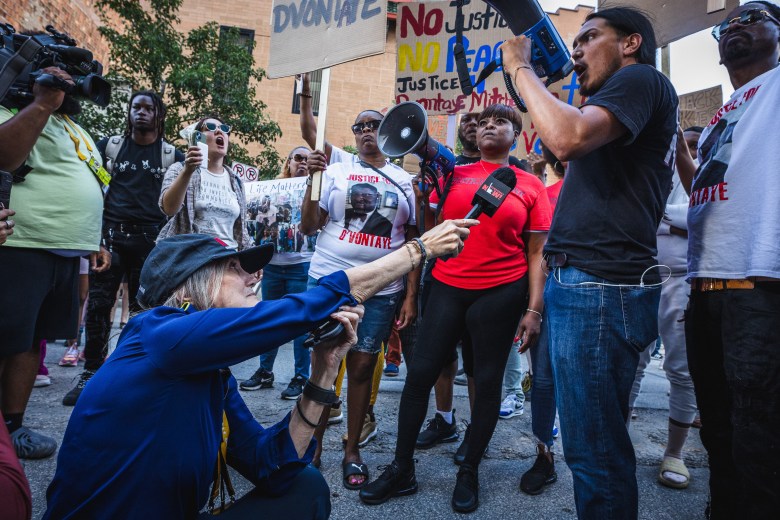 Investigative journalist Amy Goodman crouches in the street to interview a protester during a rally in Milwaukee. Goodman, wearing a navy blue long-sleeve shirt and a black baseball cap, holds a microphone up toward a person in a black t-shirt and jeans who is shouting through a blue megaphone. In the background, a diverse crowd of protesters holds signs, including one that reads 'No Justice, No Peace.' Other members of the media, including a camera operator in a lime green shirt, are visible in the busy urban setting.