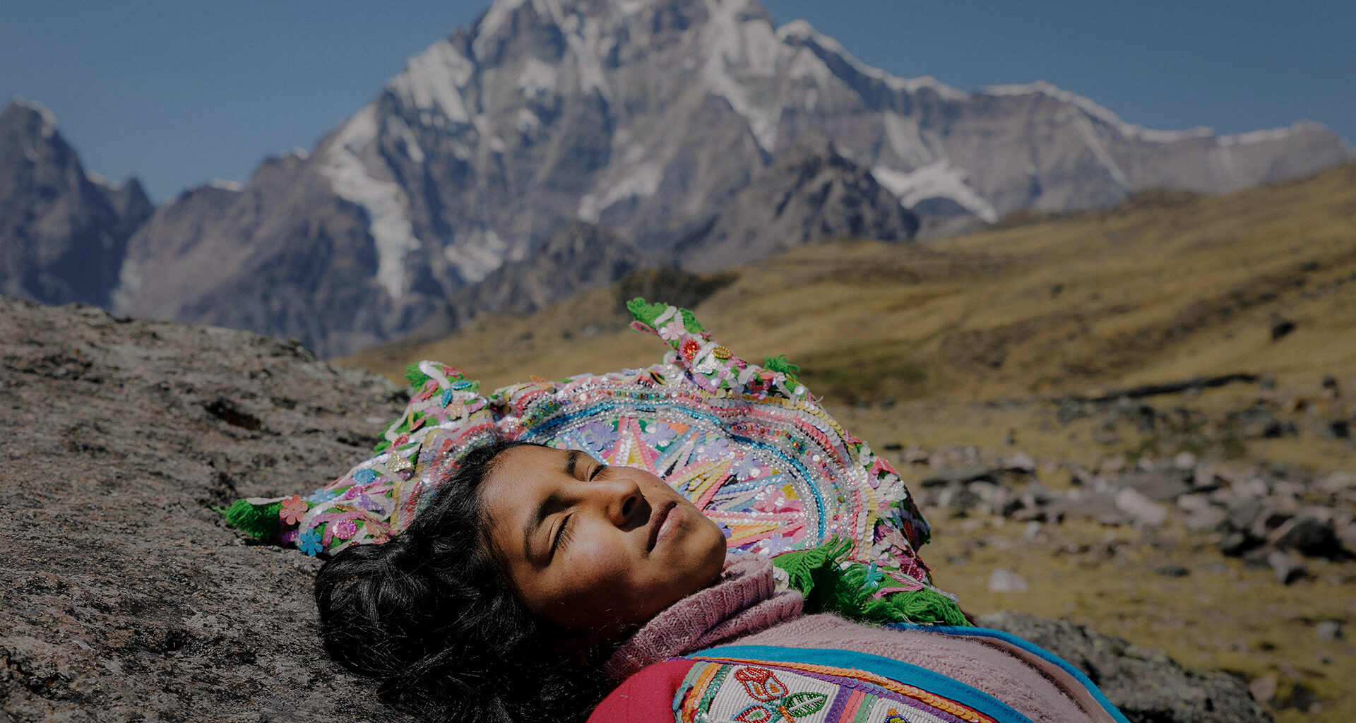 Photo of an Indigenous woman in colorful traditional clothing lying on the ground in a mountainous area. Behind her are mountains in the distance