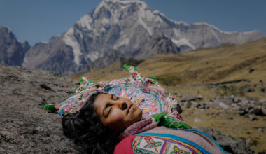Photo of an Indigenous woman in colorful traditional clothing lying on the ground in a mountainous area. Behind her are mountains in the distance