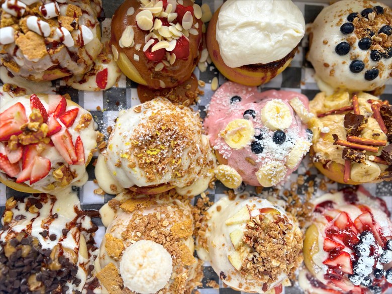 A box of twelve gourmet cinnamon rolls, each generously covered with unique frostings and varied toppings like fresh fruit, chocolate, and nuts