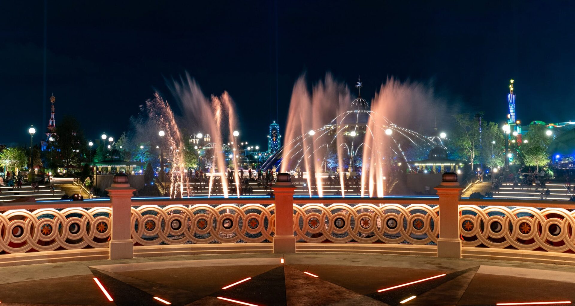 Epic Universe: Colorful fountains and lights illuminate a park at night, viewed from behind a decorative railing.