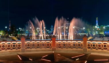 Epic Universe: Colorful fountains and lights illuminate a park at night, viewed from behind a decorative railing.